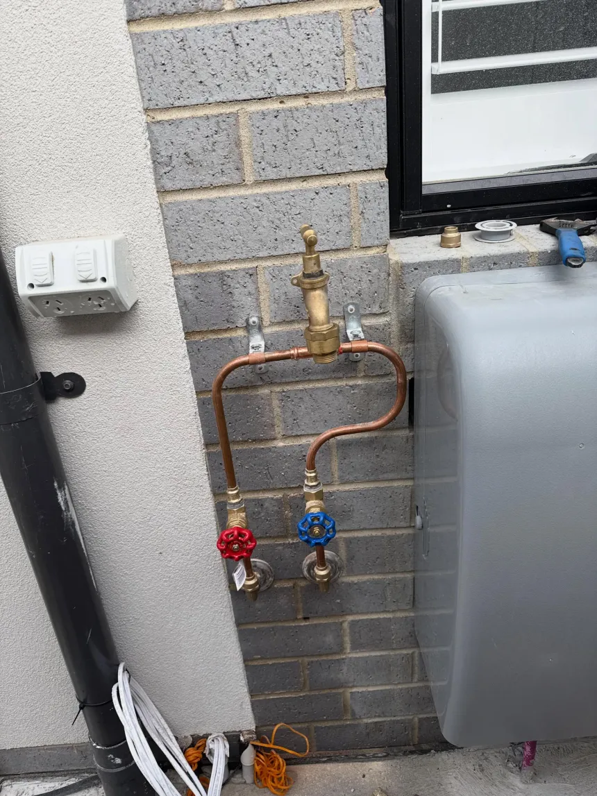 Copper pipework with a brass tempering valve above red and blue isolation valves, mounted on a brick wall beside a hot water unit